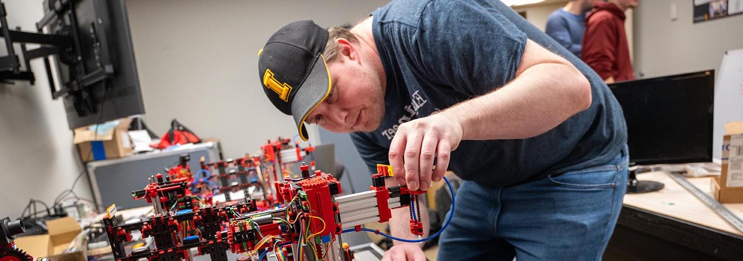 一名戴着爱达荷州棒球帽的年轻人正在调整机器人系统手臂上的一个部件，这个机器人系统模拟了机器人教室里的一个迷你工厂.